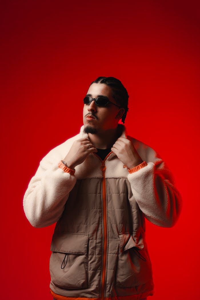 Portrait of a fashionable man with sunglasses and braided hair in a stylish jacket under red studio lighting.