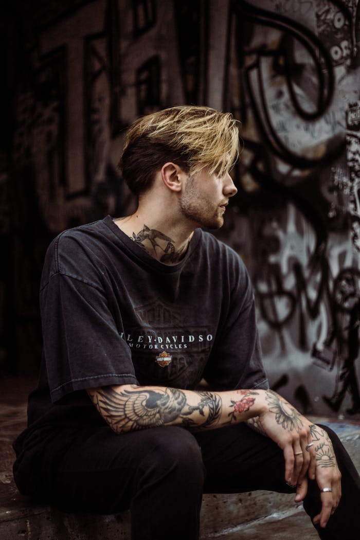 services-04 A tattooed man in a black T-shirt sits in a graffiti-covered urban environment, showcasing street style.