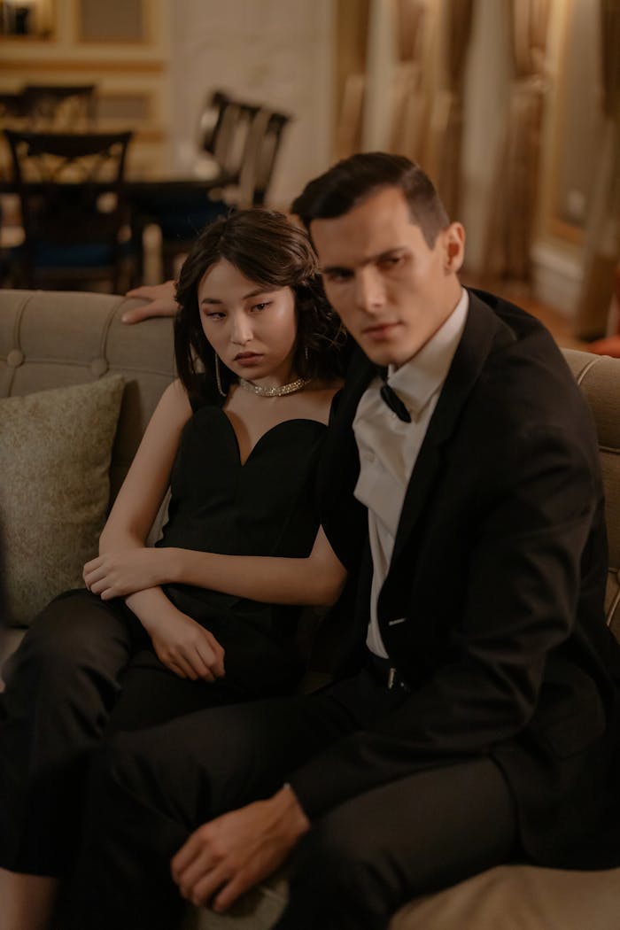 gallery-1 A couple elegantly dressed, embracing lovingly on a couch in a luxurious indoor setting.