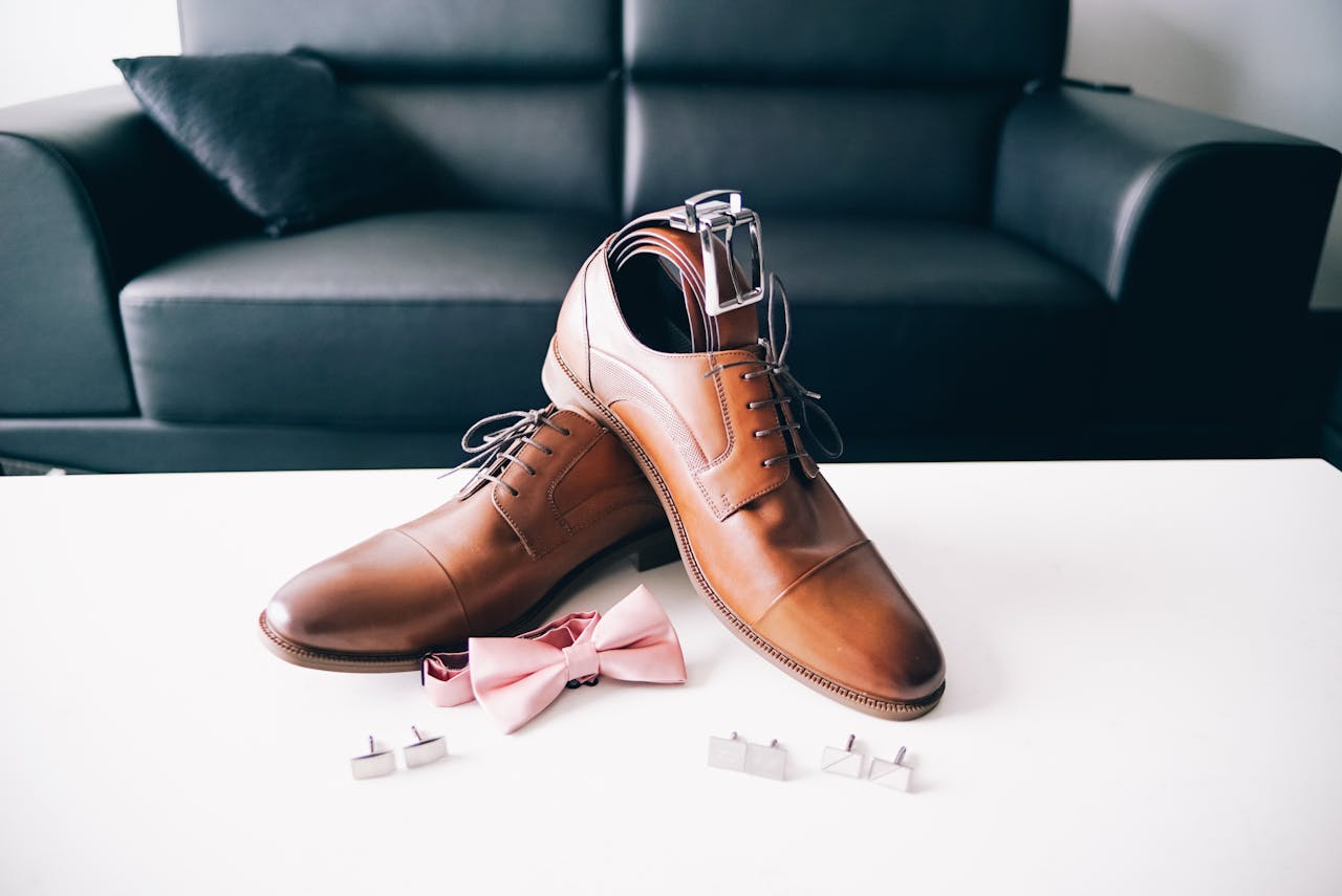 why-choose-us Stylish men's brown dress shoes with a pink bow tie and cufflinks placed on a white table, perfect for formal events.